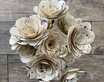 May include: A bouquet of paper flowers made from vintage sheet music. The flowers are cream-colored with black musical notes and text, arranged against a gray wooden background. The bouquet has a variety of sizes and shapes.
