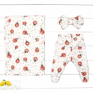 May include: A white baby outfit with a ladybug print. The outfit includes a headband, pants, and a swaddle blanket.