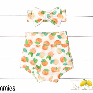 May include: White baby bloomers with a peach print and a matching peach print headband. The headband has a bow in the center. The text "Bummies" is in the lower left corner of the image. The text "Unique Little Baby Nursery & Apparel" is in the lower right corner of the image.
