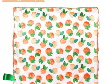 May include: A peach-colored baby blanket with a white background and a repeating pattern of peach fruit and green leaves. The blanket has a peach-colored border and a green ribbon attached to one corner.