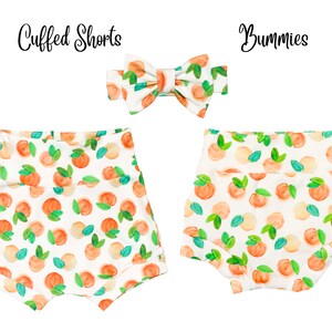 May include: A set of two peach-print baby clothes, including a pair of cuffed shorts and a bow. The shorts have a white background with peach and green watercolor-style illustrations. The bow is white with peach and green watercolor-style illustrations.
