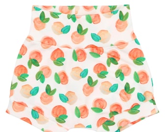 May include: White baby shorts with a peach and green print. The peaches are painted in a watercolor style.