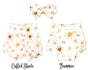 May include: A set of two white baby shorts with a sun and rainbow print. The shorts are available in two styles: cuffed shorts and bummers. A matching white headband with a sun and rainbow print is also included.