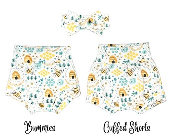 May include: A set of two baby clothes items: a pair of white cuffed shorts with a beehive and bee pattern and a white headband with a yellow bow. The text "Bummies" and "Cuffed Shorts" is printed below the items.