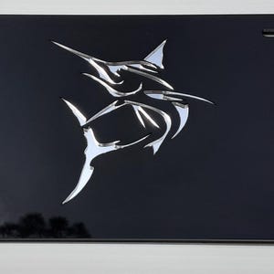 May include: Black license plate with a stylized, chrome marlin fish emblem. The plate has rounded corners and four mounting holes. The marlin design is a prominent, reflective silver against the dark background.