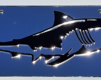 May include: A chrome license plate with a detailed black shark silhouette. The shark is depicted with open jaws and visible teeth, set against a reflective blue background. The plate has four mounting holes.