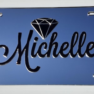 May include: A reflective license plate with the name "Michelle" in a stylish script font. A black diamond graphic is above the name. The plate has a blue background and four mounting holes.