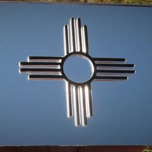May include: A shiny, chrome-like license plate featuring the Zia sun symbol, a Native American design. The plate has four mounting holes and a reflective surface. The Zia symbol is a circle with four groups of four rays extending outward.