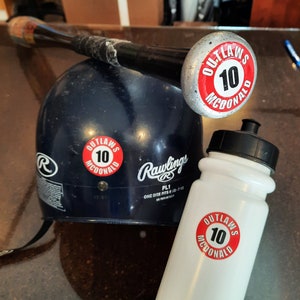 May include: A white water bottle with a red and white sticker that says "Outlaws 10 McDonald" on it. A blue baseball helmet with a Rawlings logo and a similar sticker is in the background.