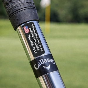 K&ouml;nnte beinhalten: Nahaufnahme eines Golfschl&auml;gers mit schwarzem Griff und silbernem Schaft. Ein schwarzer Aufkleber mit der amerikanischen Flagge, dem Namen "JUSTIN COLEMAN" und einer Telefonnummer ist angebracht. Das Callaway-Logo ist sichtbar.