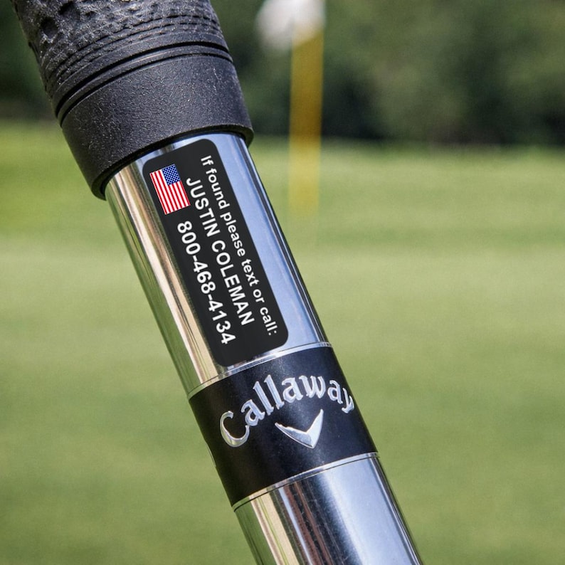 K&ouml;nnte beinhalten: Nahaufnahme eines Golfschl&auml;gers mit schwarzem Griff und silbernem Schaft. Ein schwarzer Aufkleber mit der amerikanischen Flagge, dem Namen "JUSTIN COLEMAN" und einer Telefonnummer ist angebracht. Das Callaway-Logo ist sichtbar.