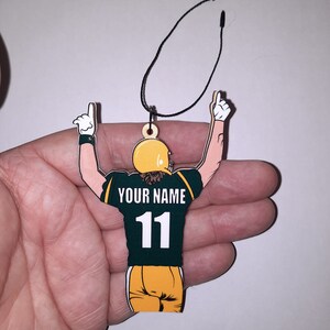 May include: Green and gold wooden ornament of a football player with arms raised in celebration. The player is wearing a jersey with the number 11 and the text "YOUR NAME".