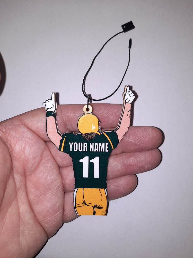 May include: Green and gold wooden ornament of a football player with arms raised in celebration. The player is wearing a jersey with the number 11 and the text "YOUR NAME".