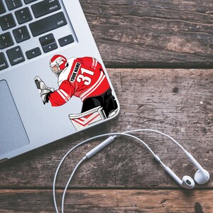 May include: A red and white hockey goalie sticker with the text "YOUR NAME" and the number "31" on the back of the jersey. The sticker is on a silver laptop.