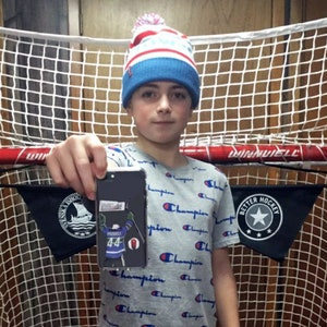 May include: A young person wearing a blue and white knit hat with a pom-pom on top is holding a black smartphone with a hockey player sticker on the back. The sticker features a hockey player wearing a blue jersey with the number 44 on it. The person is standing in front of a hockey net.