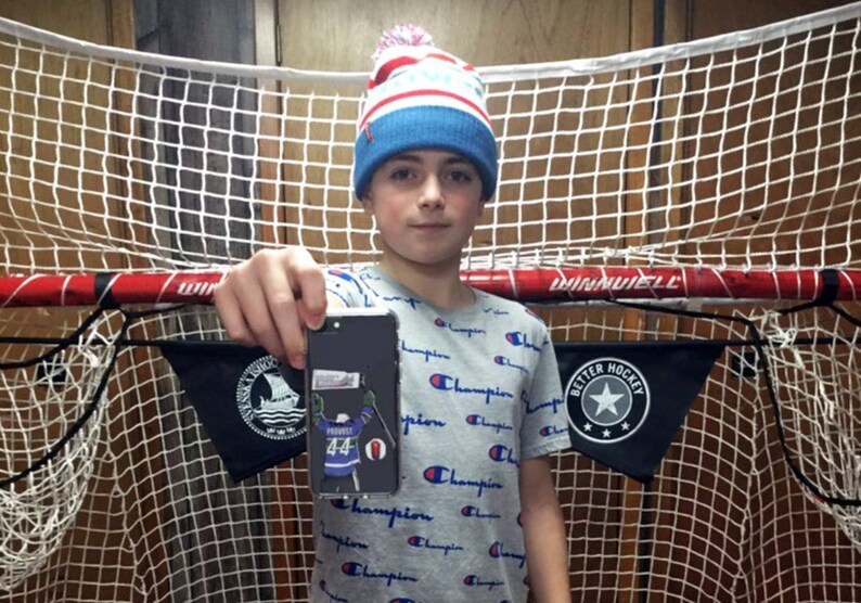 May include: A young person wearing a blue and white knit hat with a pom-pom on top is holding a black smartphone with a hockey player sticker on the back. The sticker features a hockey player wearing a blue jersey with the number 44 on it. The person is standing in front of a hockey net.
