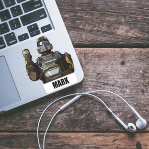 May include: A sticker featuring a cartoon illustration of a robot wearing a helmet and holding a weapon. The sticker is on a laptop and has the word "MARK" printed on it.