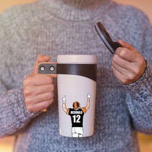 Puede incluir: Taza de viaje blanca con tapa negra y asa marr&oacute;n. La taza tiene un gr&aacute;fico de un jugador de f&uacute;tbol americano que lleva un uniforme negro y naranja con el n&uacute;mero 12 y el nombre "MCDONALD" en la espalda.