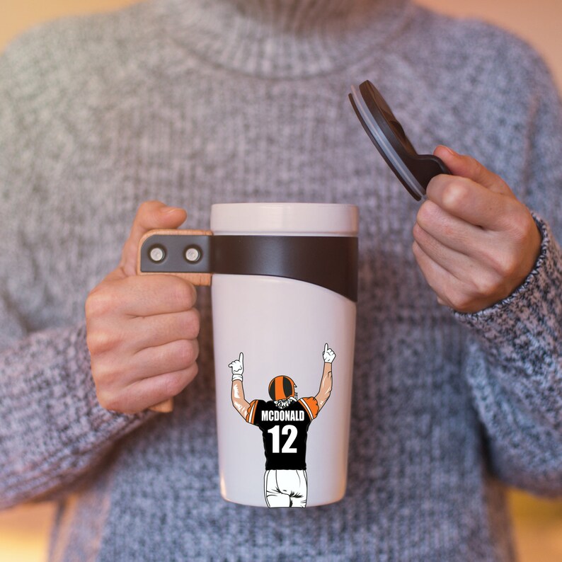 Puede incluir: Taza de viaje blanca con tapa negra y asa marr&oacute;n. La taza tiene un gr&aacute;fico de un jugador de f&uacute;tbol americano que lleva un uniforme negro y naranja con el n&uacute;mero 12 y el nombre "MCDONALD" en la espalda.