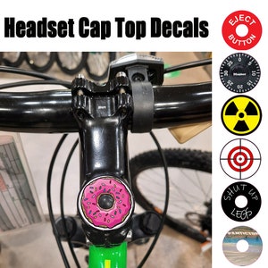 May include: A close-up of a bicycle headset cap with a pink donut sticker. The sticker has pink frosting and colorful sprinkles. The bicycle headset cap is black and has a 5mm bolt. The image also shows a variety of other stickers, including a red target, a yellow and black radiation symbol, a black and white sticker with the text "Shut Up Legs", and a red sticker with the text "Eject Button".