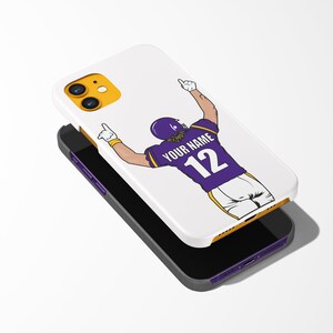 Puede incluir: Carcasa blanca para iPhone con un dise&ntilde;o morado y amarillo que presenta a un jugador de f&uacute;tbol americano celebrando un touchdown. La camiseta del jugador tiene el texto "YOUR NAME" y el n&uacute;mero "12" en la espalda.