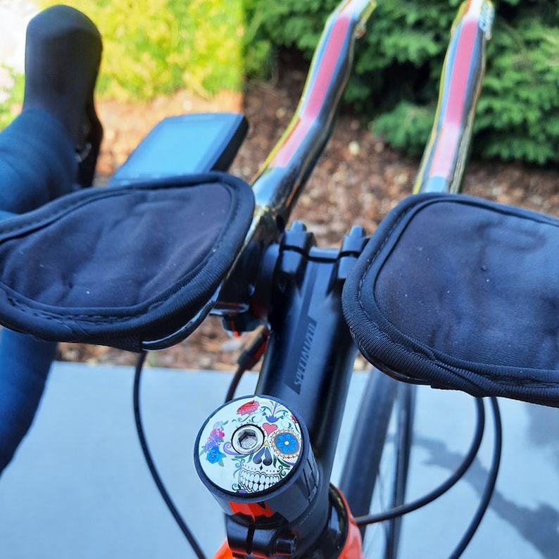 May include: A black bicycle handlebar with black handlebar grips and a colorful skull and flower design on the stem cap.