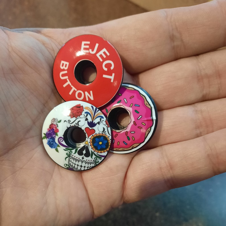 May include: Three round, colorful, printed bike caps. One is red with white lettering that says "EJECT BUTTON". One is pink with white sprinkles and a pink donut hole. One is white with a black and white skull and floral design.