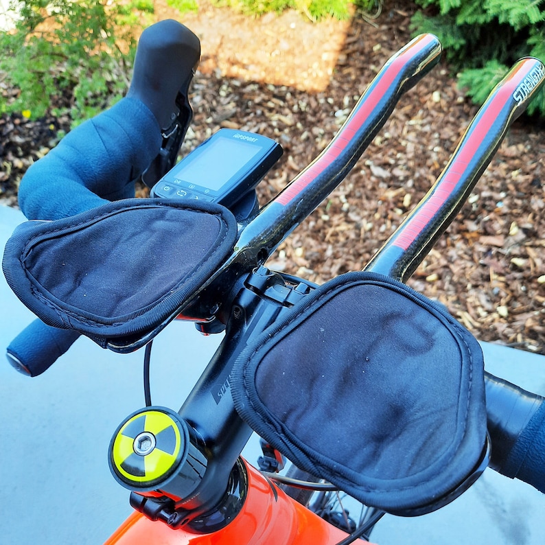 May include: A black bicycle handlebar with red accents and two black padded armrests. The handlebar has a black and yellow bike stem cap with a nuclear symbol. A black and white cycling computer is mounted on the handlebar.