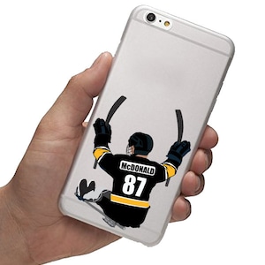 May include: A white iPhone case with a black and yellow hockey player graphic. The player is wearing a jersey with the name "MCDONALD" and the number "87".