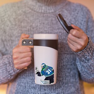 May include: White travel mug with a brown handle and a black lid. The mug features a graphic of a hockey player wearing a blue and green jersey with the number 31 and the name "McDonald" on the back.