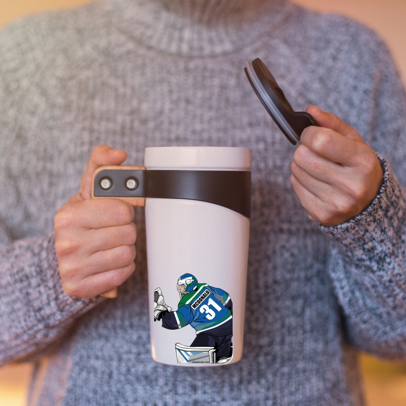 May include: White travel mug with a brown handle and a black lid. The mug features a graphic of a hockey player wearing a blue and green jersey with the number 31 and the name "McDonald" on the back.