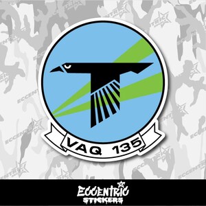May include: A sticker featuring a black bird design within a blue and green circle. The text "VAQ 135" is displayed on a white banner below the circle. The sticker has a white border and a camouflage background.