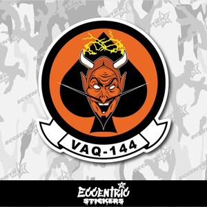 May include: A circular sticker featuring a devil character with horns, a mustache, and a sinister grin, set against a black spade shape within an orange circle. The text "VAQ-144" is displayed on a white banner below the central image.