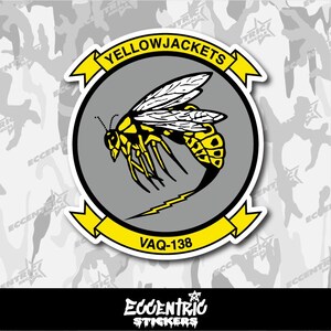 May include: A sticker featuring a stylized yellowjacket insect with black accents, set against a gray circle. The words "YELLOWJACKETS" and "VAQ-138" are displayed on yellow banners. The background is a camouflage pattern.