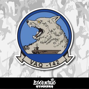 May include: A sticker featuring a wolf's head with an open mouth, set against a blue circle. Below the wolf is a ship, and a banner reads "VAQ-142". The sticker has a white border and the text "ECCENTRIC STICKERS" at the bottom.