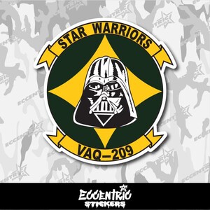 May include: A circular sticker featuring Darth Vader's helmet in black and white, set against a yellow starburst and a dark green background. The words "STAR WARRIORS" and "VAQ-209" are displayed in yellow. The sticker has a white border.