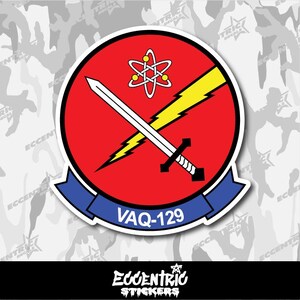 May include: A round sticker featuring a red circle with a white sword and yellow lightning bolt crossing it. Above is a white atom symbol. The sticker has a blue banner with "VAQ-129" and the text "Eccentric Stickers" at the bottom.