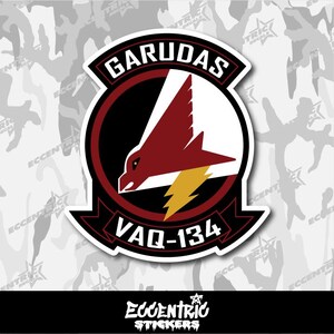 May include: A sticker featuring the "GARUDAS" emblem, a stylized eagle with a red wing and a yellow lightning bolt. The emblem is set against a black and white camouflage pattern. The text "VAQ-134" is also visible.