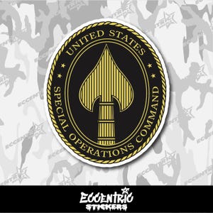 May include: A circular sticker featuring the emblem of the United States Special Operations Command. The design includes a black spade shape with yellow vertical lines, surrounded by text in a gold ring on a white background.