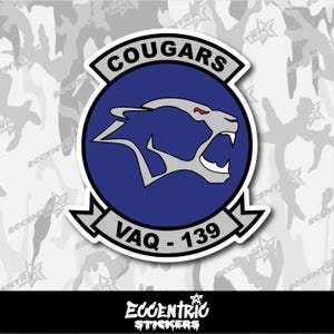 May include: A white sticker featuring a navy blue circle with a cougar head graphic. The text "COUGARS" is above the circle, and "VAQ - 139" is below. The sticker has a white border and a camouflage background.