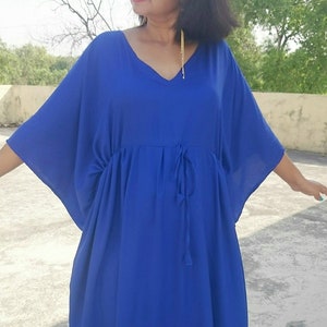 May include: A vibrant blue caftan dress with a V-neckline and a drawstring waist. The dress has wide sleeves and falls to mid-calf. The person is wearing beige sandals. The background is a concrete surface with a tree in the distance.