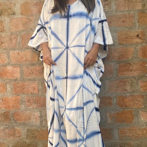 May include: A white and blue tie-dye caftan dress. The dress has a V-neck and wide sleeves. The blue dye pattern forms a geometric design across the fabric. The floor-length dress appears to be made of a lightweight material, suitable for warmer weather.