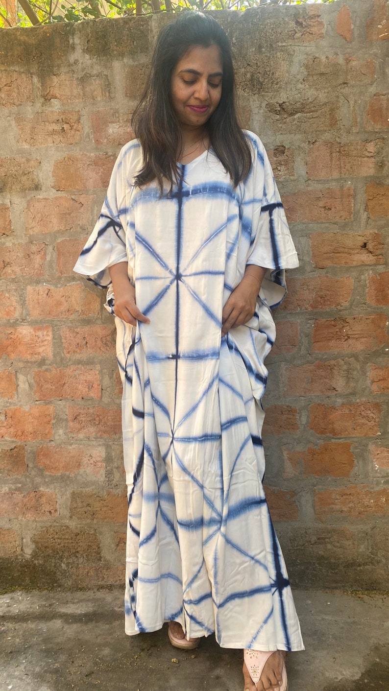 May include: A white and blue tie-dye caftan dress. The dress has a V-neck and wide sleeves. The blue dye pattern forms a geometric design across the fabric. The floor-length dress appears to be made of a lightweight material, suitable for warmer weather.