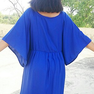 May include: A vibrant blue dress with wide, flowing sleeves and a cinched waist. The dress is made of a lightweight fabric and falls to the mid-calf. The model's back is to the camera, showcasing the dress's design and silhouette.