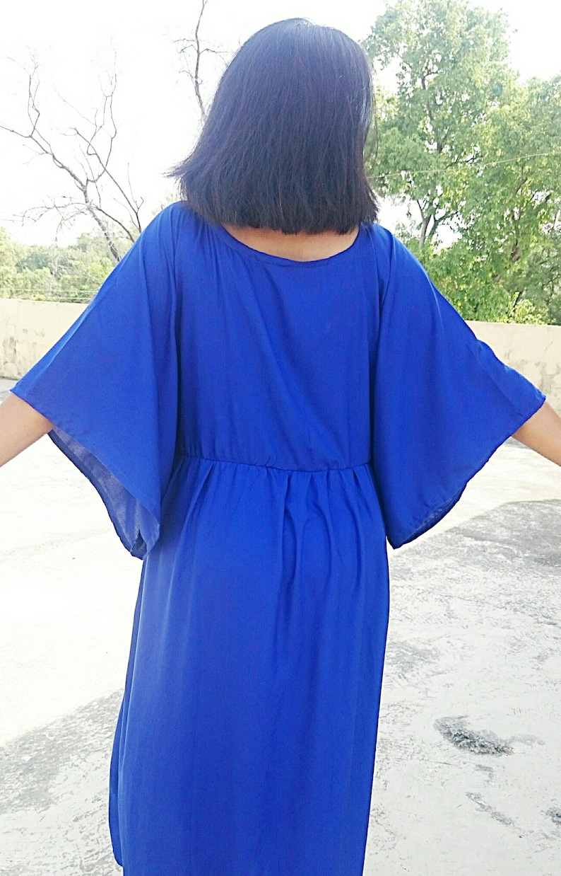 May include: A vibrant blue dress with wide, flowing sleeves and a cinched waist. The dress is made of a lightweight fabric and falls to the mid-calf. The model's back is to the camera, showcasing the dress's design and silhouette.