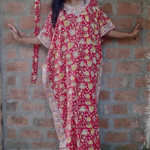 May include: A red and yellow floral print kaftan with white trim. The kaftan has a long, flowing silhouette and is made of a lightweight fabric.