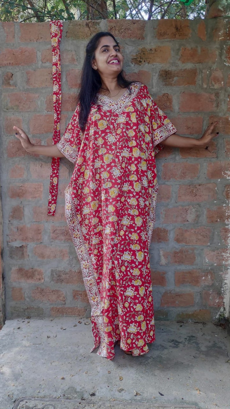 May include: A red and yellow floral print kaftan with white trim. The kaftan has a long, flowing silhouette and is made of a lightweight fabric.