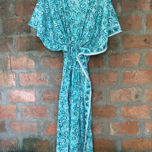 May include: A teal blue kaftan with a white floral print. The kaftan has a V-neck and short sleeves. It is made of a lightweight fabric and has a tie at the waist.