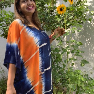 May include: A woman wearing a blue and orange tie-dye kaftan with a V-neck and short sleeves. She is standing in a garden with sunflowers in the background.