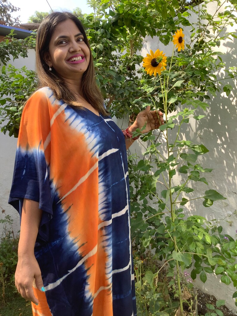 May include: A woman wearing a blue and orange tie-dye kaftan with a V-neck and short sleeves. She is standing in a garden with sunflowers in the background.
